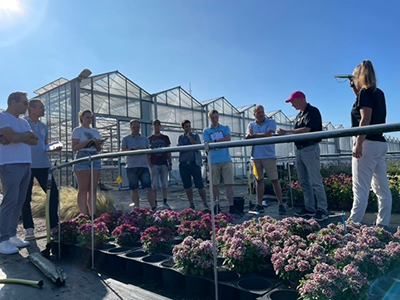 Glass greenhouses are the backdrop to several grower customers listening to a presenter in a trials display on black cloth.