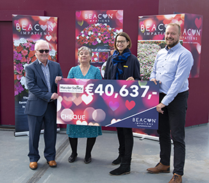 Three people hold an oversized check in Euros in front of Beacon Impatiens signage.